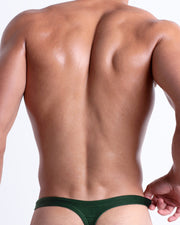 Back view of a model wearing CASINO ROYALE (GREEN) men’s beach swim thong  in dark green color. Inspired by actor Daniel Craig's iconic blue swim trunks worn in the 2006 film Casino Royale, is designed by BANG! Clothes in Miami.
