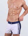 Frontal view of a male model wearing the CASINO ROYALE (WHITE + NAVY BLUE) Beach Shorts for men by BANG! Miami. These modern-fit swim shorts feature a striking white body with a navy blue contrast waistband, gold-tone dual button accents, and a sleek, contoured fit designed for beach and poolside elegance.