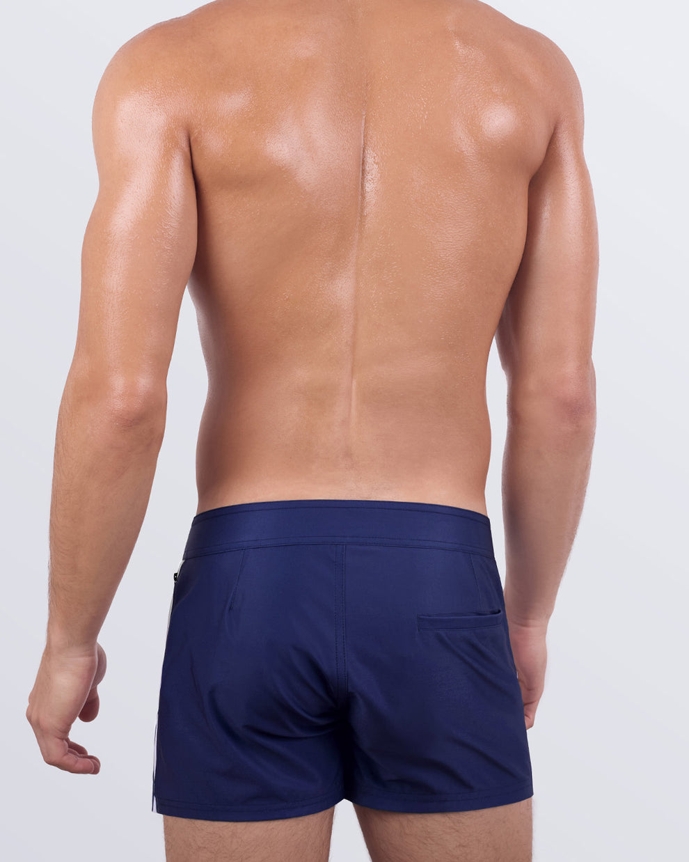 Back view of a male model showcasing the CASINO ROYALE (WHITE + NAVY BLUE) Beach Shorts by BANG! Miami. These tailored beach shorts feature a minimalist back pocket design, a seamless navy blue waistband, and a comfortable stretch fabric for unrestricted movement.