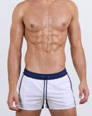 Frontal view of a male model wearing the CASINO ROYALE (WHITE + NAVY BLUE) Show Shorts for men by BANG! Miami. These stylish and ultra-modern beach shorts feature a white base with navy blue accents, a flat elastic waistband with drawstring fastening, and a tailored quad-friendly fit, perfect for beach days and resort lounging.