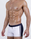 Side front view of the CASINO ROYALE Swim Trunks in White + Navy Blue – sleek square-cut silhouette with a contrast waistband and minimalist side stripe.