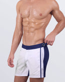 Angled front view of a male model wearing the Casino Royale (White + Navy Blue) Tailored Shorts. The adjustable side buckles allow for a customized fit, while the high-quality quick-dry fabric ensures all-day comfort by the water.