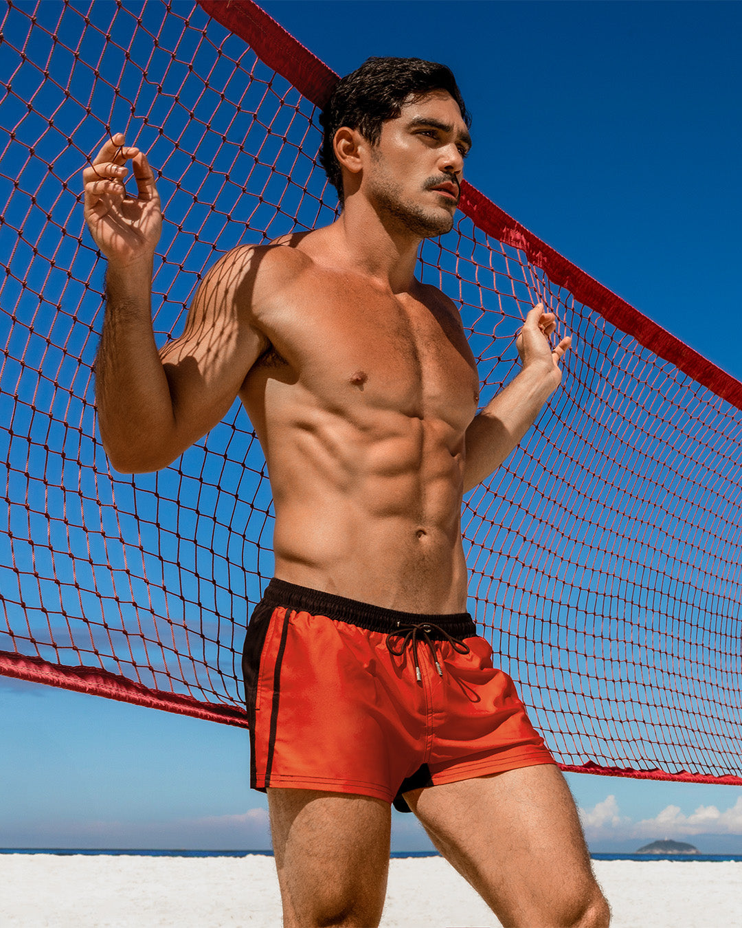DC2 Casino Royale (Rapid Red) Show Shorts — model wearing bright red beach shorts with black trim, posed against a volleyball net on a sunny beach.