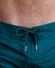 Close-up view of inseam and details of CHIC TEAL beach shorts for men, with a teal color cord and custom branded silver cord-ends, and matching custom eyelet trims in black. 