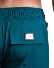 Close-up view of the CHIC TEAL men’s Flex Boardshorts back pocket, showing custom branded silver metal logo.