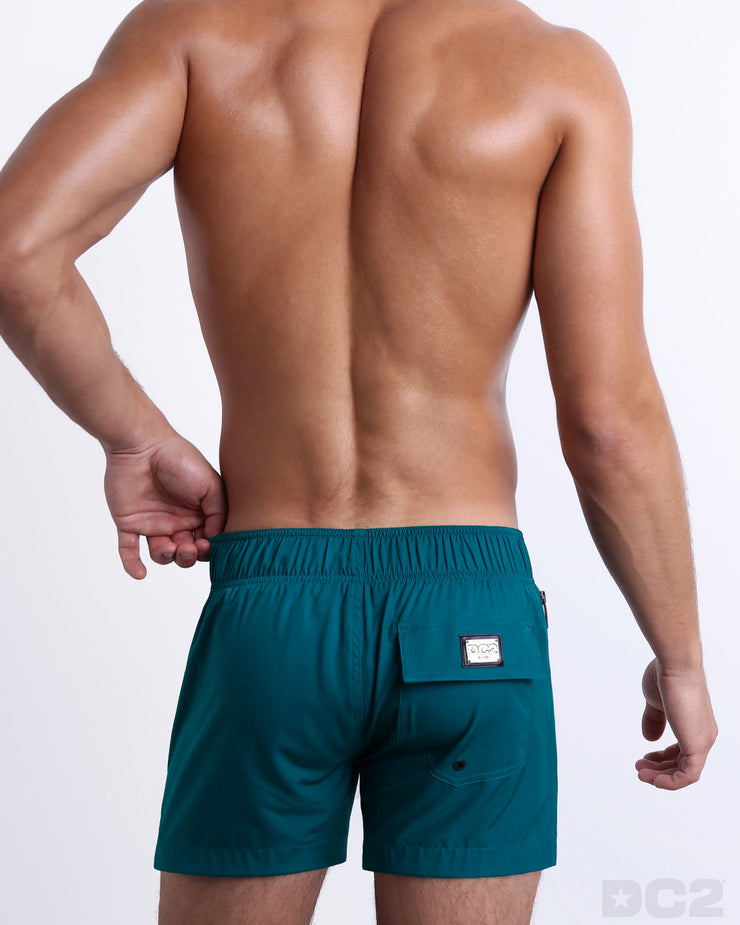 Back view of a male model wearing men’s CHIC TEAL Flex Shorts swimsuits in a solid dark teal color, complete the back pockets, made by DC2 a capsule brand by BANG! Clothes in Miami.
