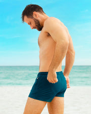 Side view of a model on the beach in CHIC TEAL Poolside Shorts by DC2 Miami. These versatile swim shorts feature a sleek fit and premium design, perfect for beach days and poolside relaxation.