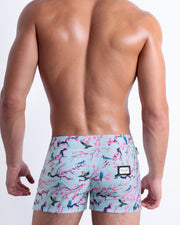 Back view of a male model wearing men’s Summer CLOSE TO YOU Beach Shorts in a light blue color with an exotic birds print, complete with a back pocket, designed by DC2.