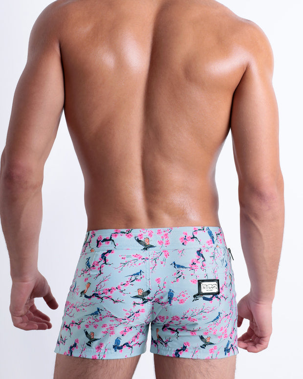 Back view of a male model wearing men’s Summer CLOSE TO YOU Beach Shorts in a light blue color with an exotic birds print, complete with a back pocket, designed by DC2.