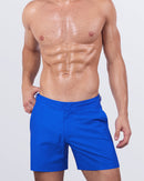 A male model wears Club Blue tailored swimming shorts for men, featuring a fitted cut, adjustable side buckles, and a hidden snap button for a sleek beachwear look.