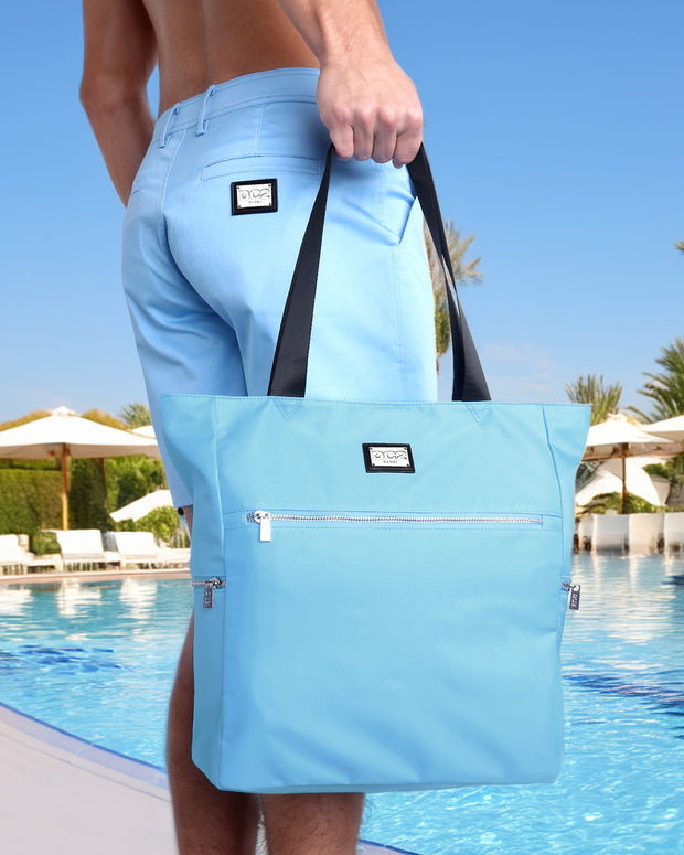 The model is poolside holding the COASTAL BLUE Poolside Tote Bag by DC2 Miami. The tote bag features a vibrant blue color, matching the BREEZY BLUE Street Shorts, with black handles and a front zippered pocket. The backdrop of a clear blue sky, palm trees, and white poolside loungers adds to the summery, beach-ready vibe of the image, highlighting the bag&