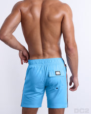 Back view of male model wearing men’s COASTAL BLUE beach Flex Boardshorts swimming shorts. In a solid blue color with side stripes in orange and dark blue colors, complete with a back pocket, designed by DC2 a capsule brand by BANG! Clothes based in Miami.