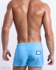 Back view of male model wearing men’s COASTAL BLUE beach Poolside Shorts swimsuits in a solid light blue color with orange and dark blue side stripes, complete the back zippered pocket, made by DC2 a capsule brand by BANG! Clothes in Miami.