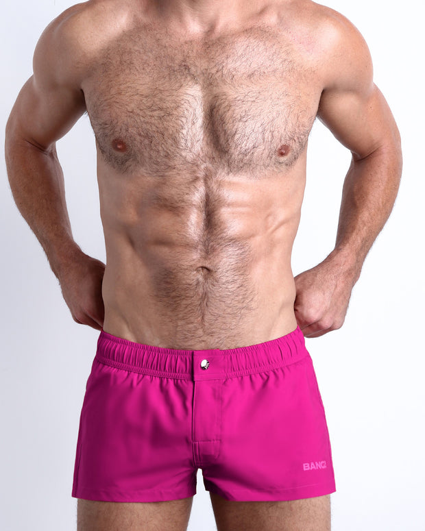 Frontal view of a sexy male model wearing the CONFESS MAGENTA men’s square leg swim trunks in a solid magenta color by the Bang! Menswear brand from Miami.