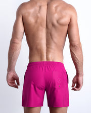 Back view of male model wearing the CONFESS MAGENTA beach trunks for men by BANG! Miami in a hot pink color with a back pocket.
