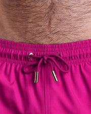 Close-up view of inseam and details of CONFESS MAGENTA swimsuit for men, with a magenta color cord and custom branded golden cord-ends, and matching custom eyelet trims in gold. 
