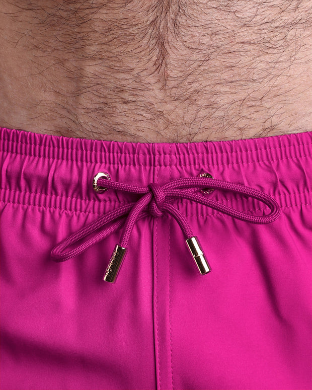 Close-up view of the CONFESS MAGENTA men’s summer shorts, showing hot pink cord with custom branded golden cord ends, and matching custom eyelet trims in gold.