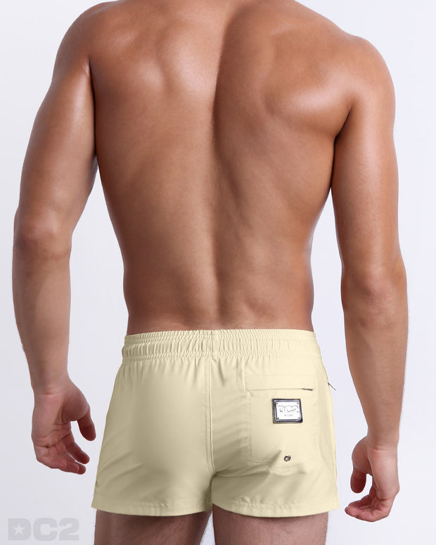 Male model wearing men’s CREAM FIELDS Summer Poolside Shorts swimsuit in a solid pale yellow color, complete with a back metallic zippered-pocket, designed by DC2 a capsule brand by BANG! Clothes based in Miami.