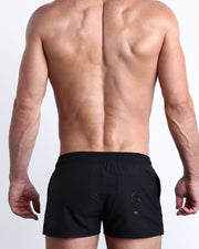 Back view of a male model wearing men’s beach trunks by BANG! menswear Miami in dark black color.