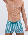 Frontal view of a masculine model wearing the FAST LINES (PACIFIC GREEN) beach shorts for men by DC2 Miami, a premium swimwear brand. These modern-fit swimming shorts feature a striking pacific green and white striped design, offering a sleek, tailored look perfect for beach days, poolside lounging, and summer getaways.