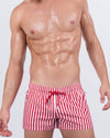Model wearing FAST LINES (RAPID RED) poolside shorts in bold red and white vertical stripes. Athletic fit swimwear with adjustable drawstring waistband, made for standout beach looks.