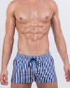 Model wearing FAST LINES (NAVY BLUE) poolside shorts in a bold navy and white vertical stripe print. Modern resortwear designed for standout beachside moments with quad-accentuating fit and metallic drawcord tips.
