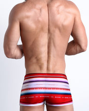 Back view of a Male model wearing swim trunks for men featuring red, blue, purple, white colors inspired by the famous 1976 swimsuit poster of Farrah Fawcettby the Bang! Clothes brand of men's beachwear.