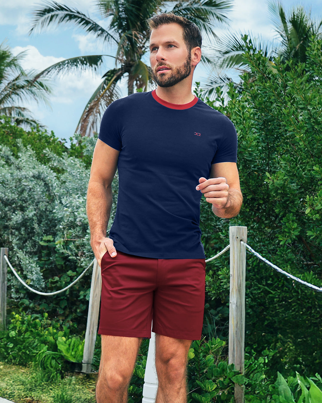 Model wearing DC2 Fontaine Blue Pima Cotton T-Shirt and Under My Umbrella Street Shorts, walking along a tropical path, showcasing a bold navy and red resort look.