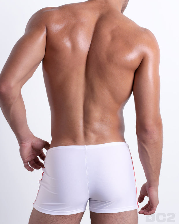 Back view of male model wearing the FORZA WHITE beach swimming bottoms for men in a solid white color with side green and red stripes, designed by DC2.