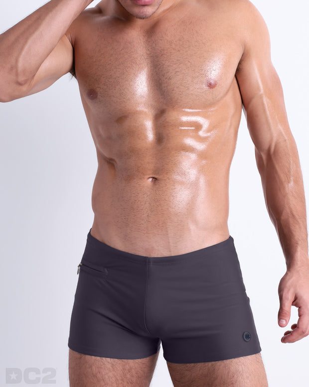 Frontal view of a sexy male model wearing men’s swimsuit with mini pocketsin a solid dark grey color by DC2, a men&