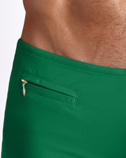 Close-up view of the GREEN RUSH Swim Trunks men’s swimsuit in a jade green color with a mini zippered pocket and white internal drawstring cord showing custom branded golden buttons by BANG! clothing brand.