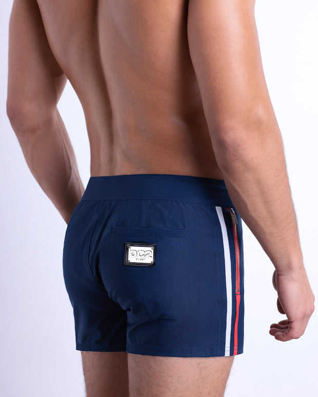 Back view of a male model wearing men’s Summer IMPERIAL BLUE Beach Shorts in a solid dark blue color with red and white side stripes, complete the back pockets, made by DC2 a capsule brand by BANG! Clothes in Miami.