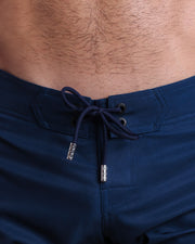 Close-up view of men’s summer long boardshorts by DC2 clothing brand, showing dark navy blue color cord with custom-branded silver cord ends, and matching custom eyelet trims in black.