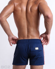 Back view of a male model wearing men’s IMPERIAL BLUE Flex Shorts swimsuits in a solid navy blue color with white and red side stripes, complete the back pockets, made by DC2 a capsule brand by BANG! Clothes in Miami.