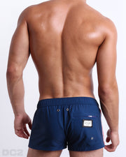 Back view of male model wearing men’s IMPERIAL BLUE beach Mini Shorts swimsuits in a solid dark blue color with side stripes in red and white colors, complete with a back pocket, designed by DC2 a capsule brand by BANG! Clothes based in Miami.