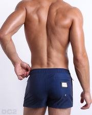 Back view of male model wearing men’s IMPERIAL BLUE beach Poolside Shorts swimsuits in a solid navy blue color with white and red side stripes, complete the back zippered pocket, made by DC2 a capsule brand by BANG! Clothes in Miami.