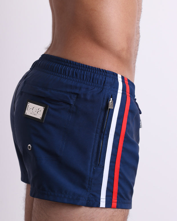 Side view of the IMPERIAL BLUE for men’s summer Poolside Shorts with dual zippered pockets. The shorts are in a solid dark blue color with side white and red stripes for men made by DC2 a brand based in Miami.