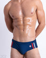 Front view of model wearing the IMPERIAL BLUE men’s bikini-style Swim Brief, premium swimwear in a solid navy blue color with white and red stripes on the sides for men. These high-quality swimwear are by DC2, a men’s beachwear brand from Miami.