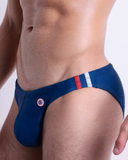 Side view of muscular male model wearing IMPERIAL BLUE Summer Swim Mini Brief. This swimsuit in dark blue color with stylish white and red colored stripes for men made by DC2 a brand based in Miami.