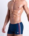 Side view of muscular male model wearing IMPERIAL BLUE Summer Swim Trunks. This swimsuit in dark blue color with stylish white and red colored stripes for men made by DC2 a brand based in Miami.