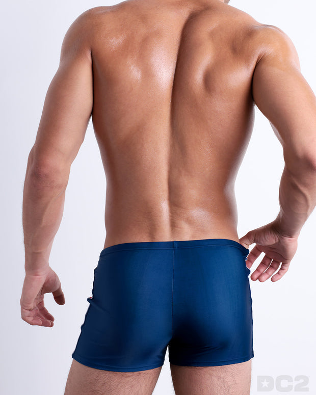 Back view of male model wearing the IMPERIAL BLUE beach swimming bottoms for men in a solid blue color with side white and red stripes, designed by DC2.