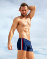 Male model wearing IMPERIAL BLUE Swim Trunks by DC2 Miami, featuring bold red and white side stripes, standing on a sandy beach. Stylish fitted men's swimwear for beach and summer fashion.