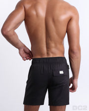 Back view of male model wearing men’s JET BLACK beach Flex Boardshorts swimming shorts. In a solid black color, complete with a back pocket, designed by DC2 a capsule brand by BANG! Clothes based in Miami.