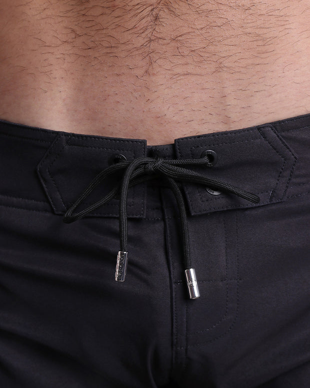 Close-up view of inseam and details of JET BLACK beach shorts for men, with a black color cord and custom branded silver cord-ends, and matching custom eyelet trims in black. 