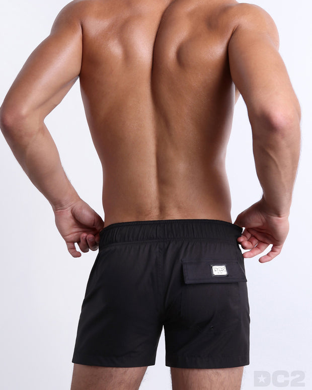 Back view of a male model wearing men’s JET BLACK Flex Shorts swimsuits in a solid black color, complete the back pockets, made by DC2 a capsule brand by BANG! Clothes in Miami.
