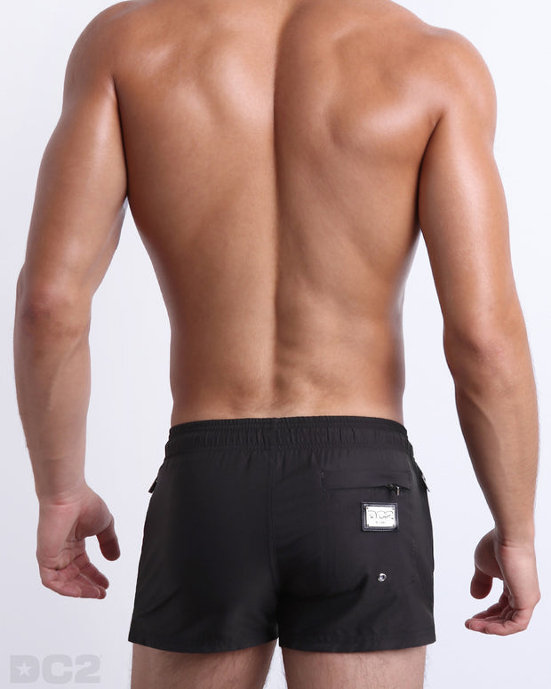 Male model wearing men’s JET BLACK Summer Poolside Shorts swimsuit in a solid black color, complete with a back metallic zippered-pocket, designed by DC2 a capsule brand by BANG! Clothes based in Miami.