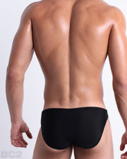Back view of male model wearing the JET BLACK beach mini-briefs for men by BANG! Miami in a solid black color.