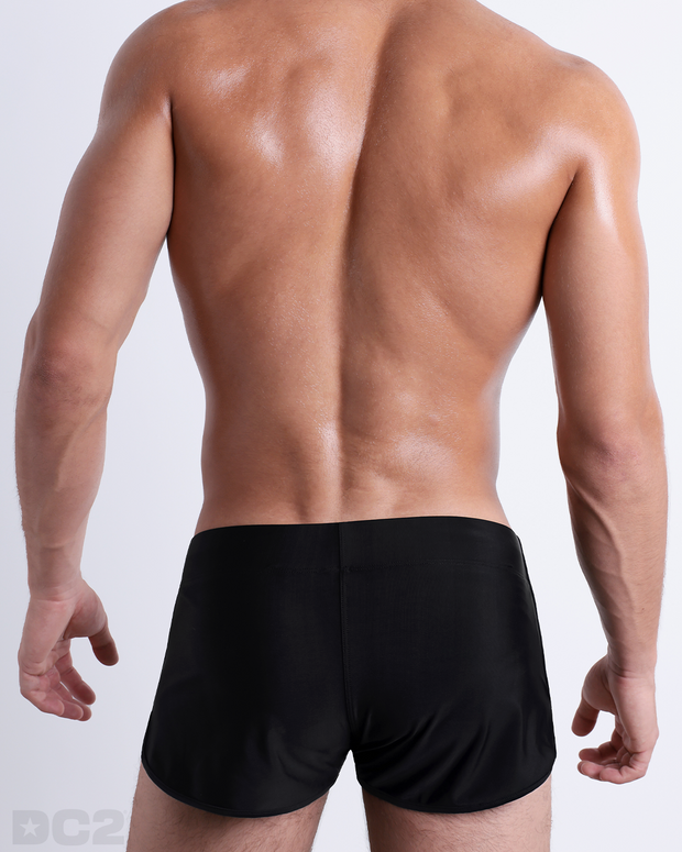 Back view of male model wearing the JET BLACK beach Swim Shorts for men by BANG! Miami in a solid black color.