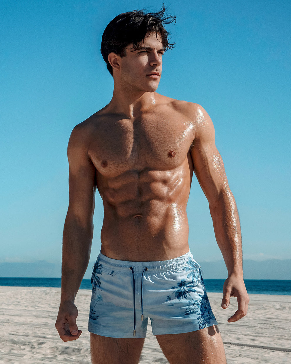 DC2 La Isla Bonita Poolside Shorts — model standing on a sunny beach wearing light blue tropical-print shorts, highlighting the tailored fit, coastal-inspired design, and vacation-ready aesthetic.