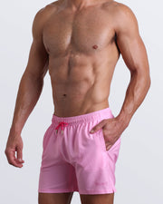 Side view of a masculine model wearing men’s boardshorts in pink with official logo of BANG! Brand in darker pink shade.